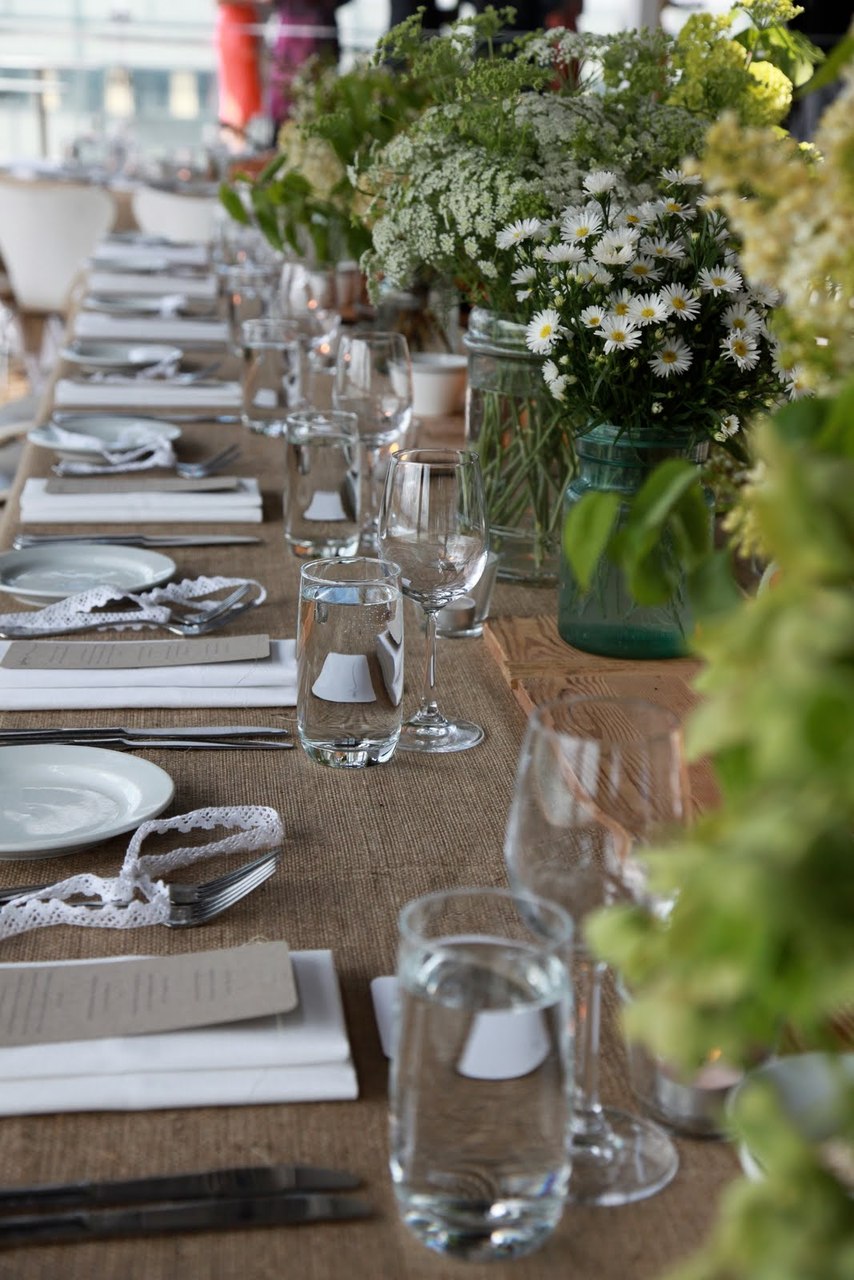 Long banquet table set with white plates, wine and water glasses, cutlery, and glass jars of white wildflowers, in a bright room