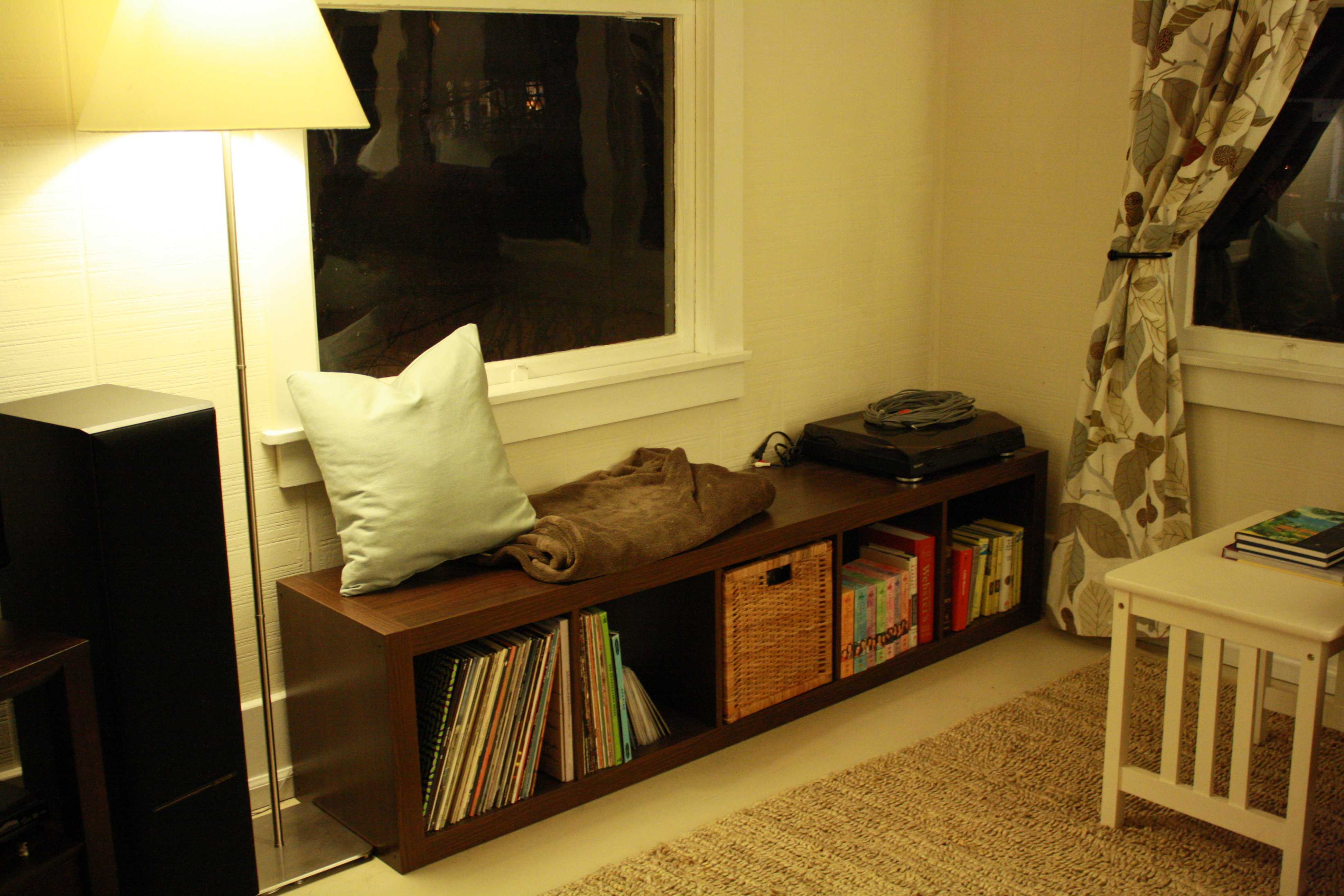 Low wooden shelving unit with books, a basket, and a cushion beneath a window, warm lamp lighting, cozy mood